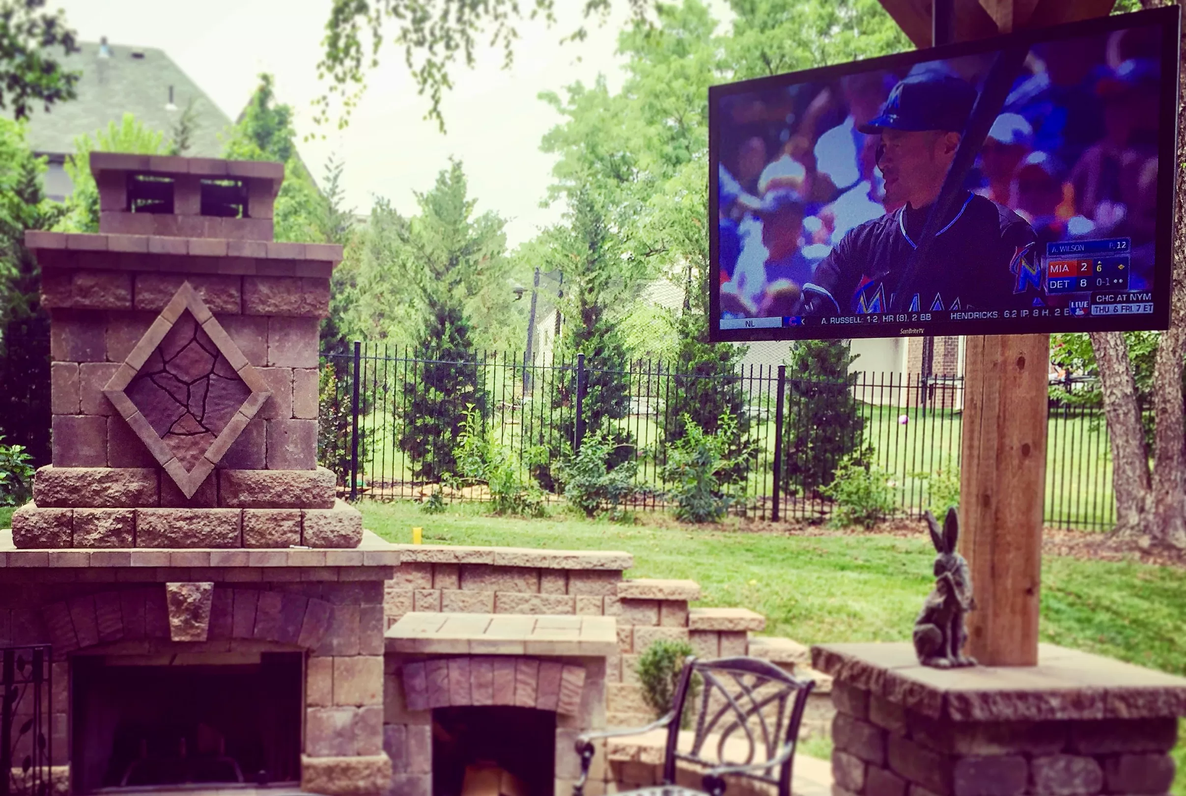 Blog 2 Outdoor television mounted on a patio post near a backyard fireplace and seating area showing a baseball game.